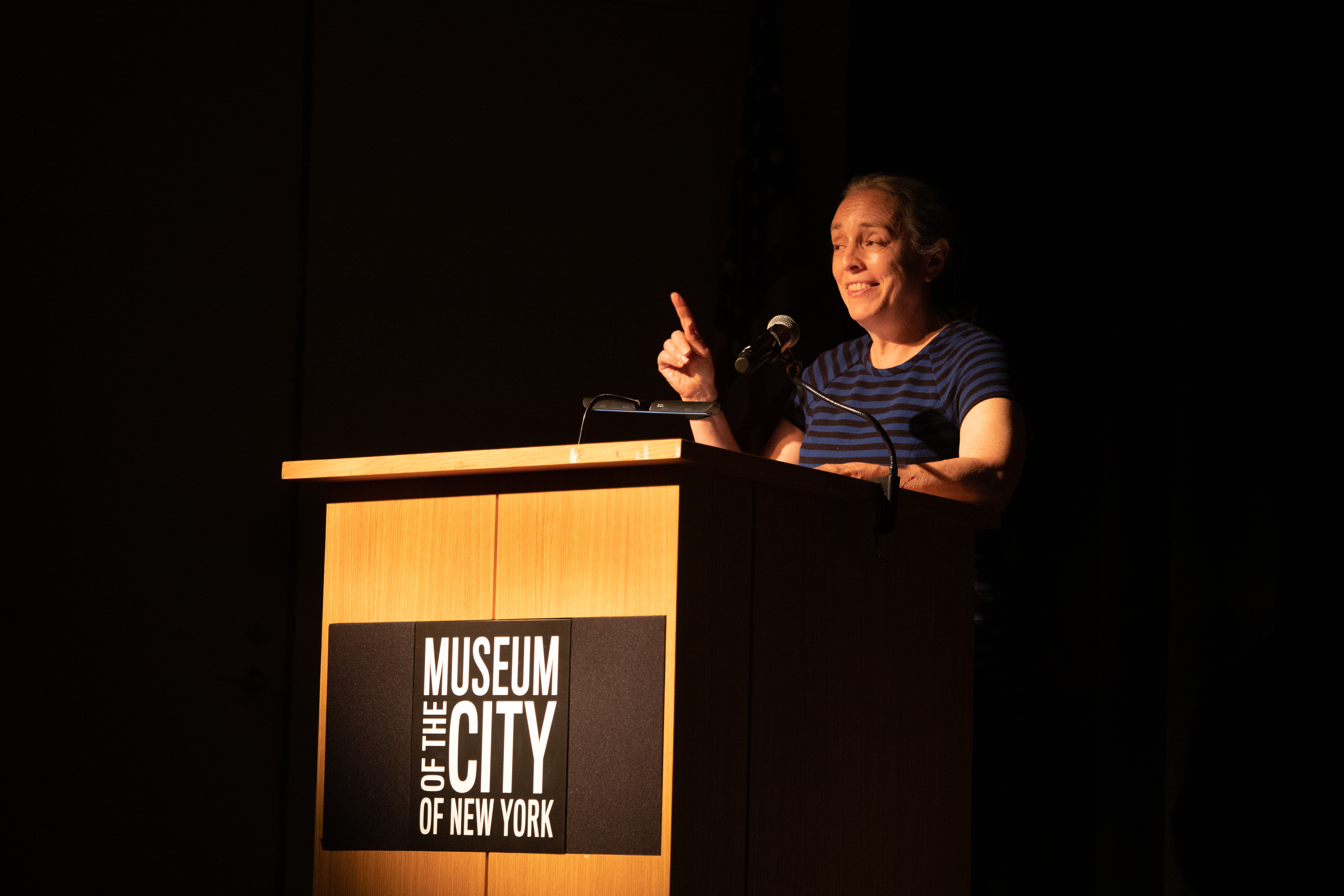 Image of woman in front of a podium labeled "Museum of the City of New York". The background is black.